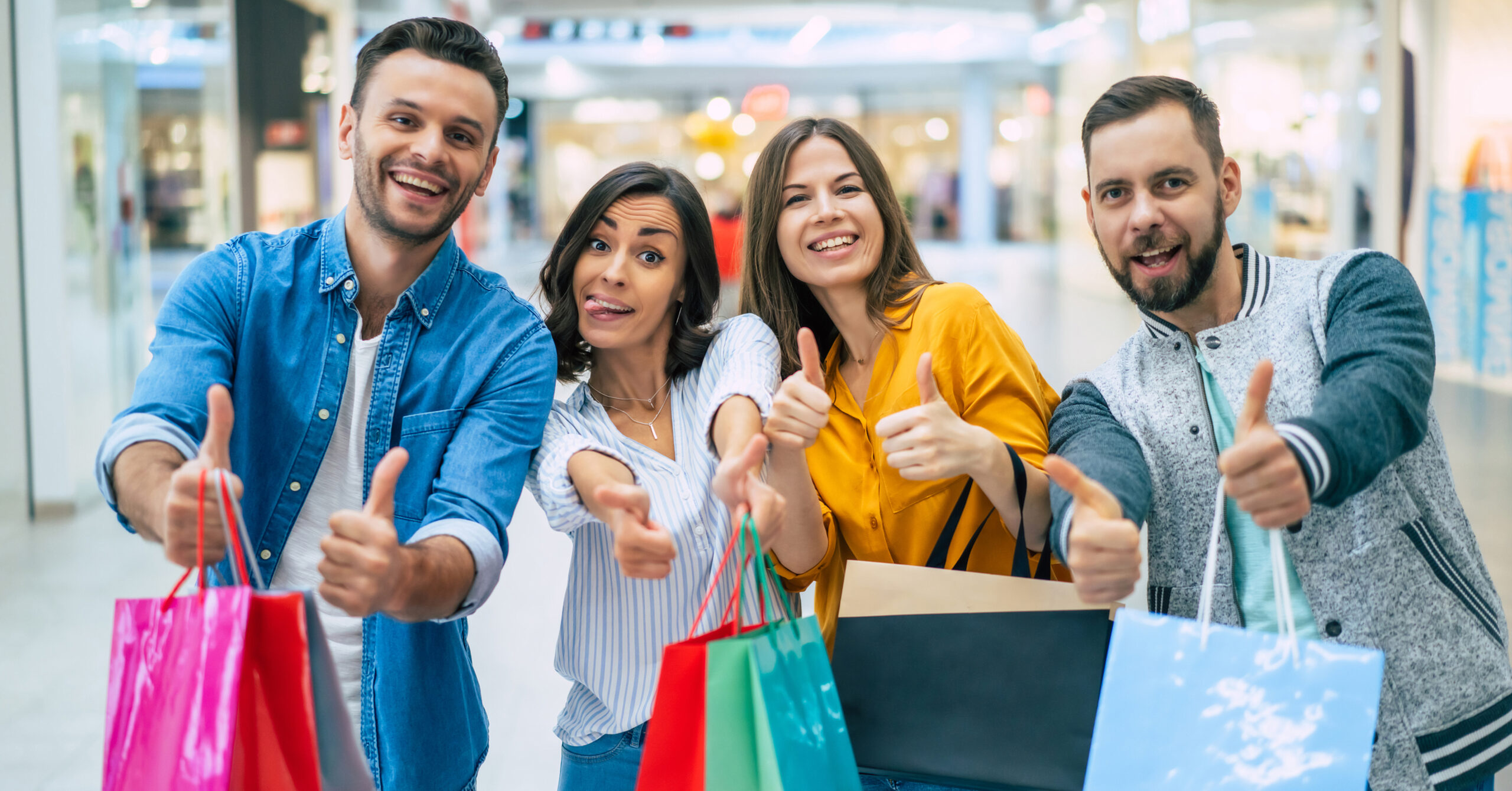 Happy company of smiling excited beautiful stylish friends with colorful paper bags are having fun and showing thumbs up on camera while shopping in the mall.